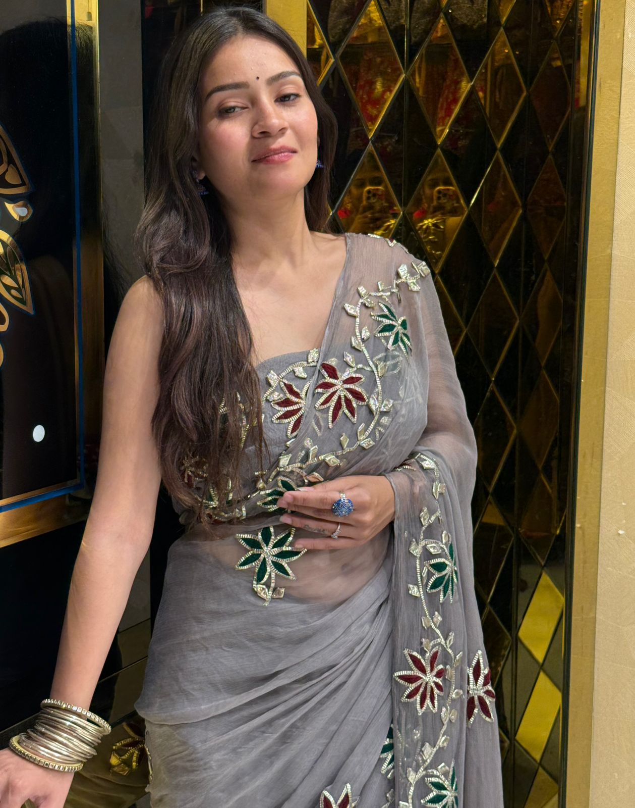 Woman in a gray saree with floral embroidery against a decorative wall.